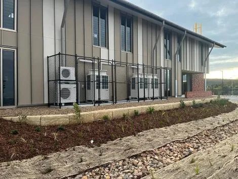 Black chain-wire cages around air conditioning units beside a new commercial building with a landscaped swale