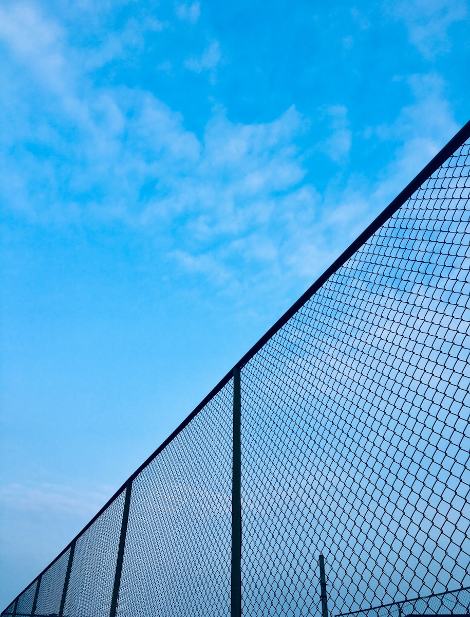 Low-angle view of a dark chain-wire fence line against a bright blue sky with light clouds